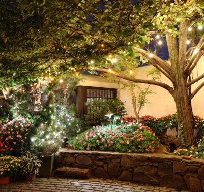 Giardino fiorito illuminato da luci decorative serali, con alberi, aiuole colorate e atmosfera accogliente.