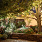 Giardino fiorito illuminato da luci decorative serali, con alberi, aiuole colorate e atmosfera accogliente.
